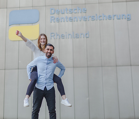 Ein junger Mann und eine junge Frau vor dem Logo der Deutschen Rentenversicherung Rheinland. Er trägt sie Huckepack. Beide strahlen freudig in die Kamera.
